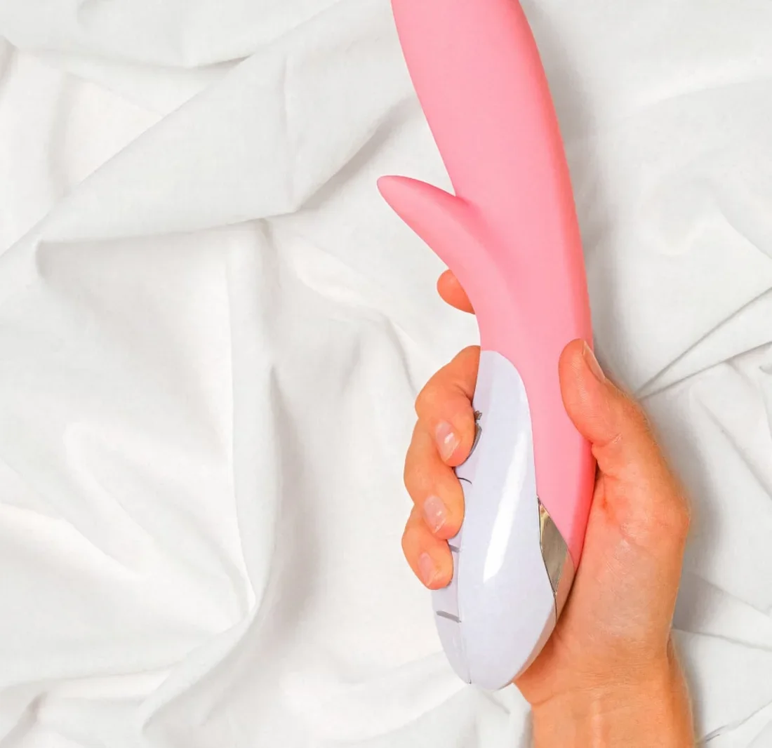 Vibrador de silicona rosa sostenido en la mano sobre sábanas blancas